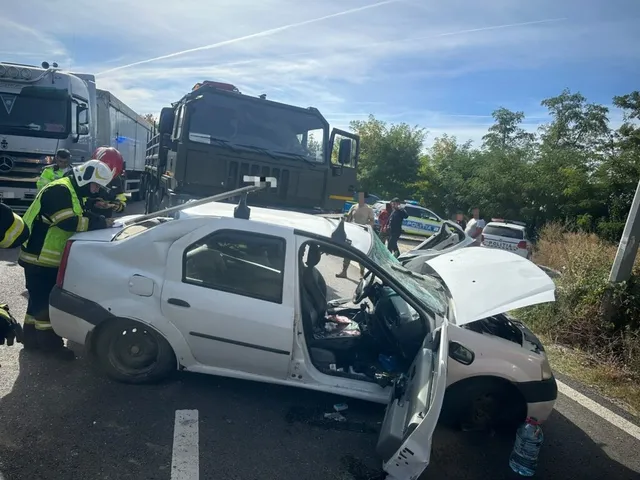 Un autoturism alb cu portierele deschise și capota ridicată stă pe asfalt lângă un camion negru și un vehicul de poliție.
