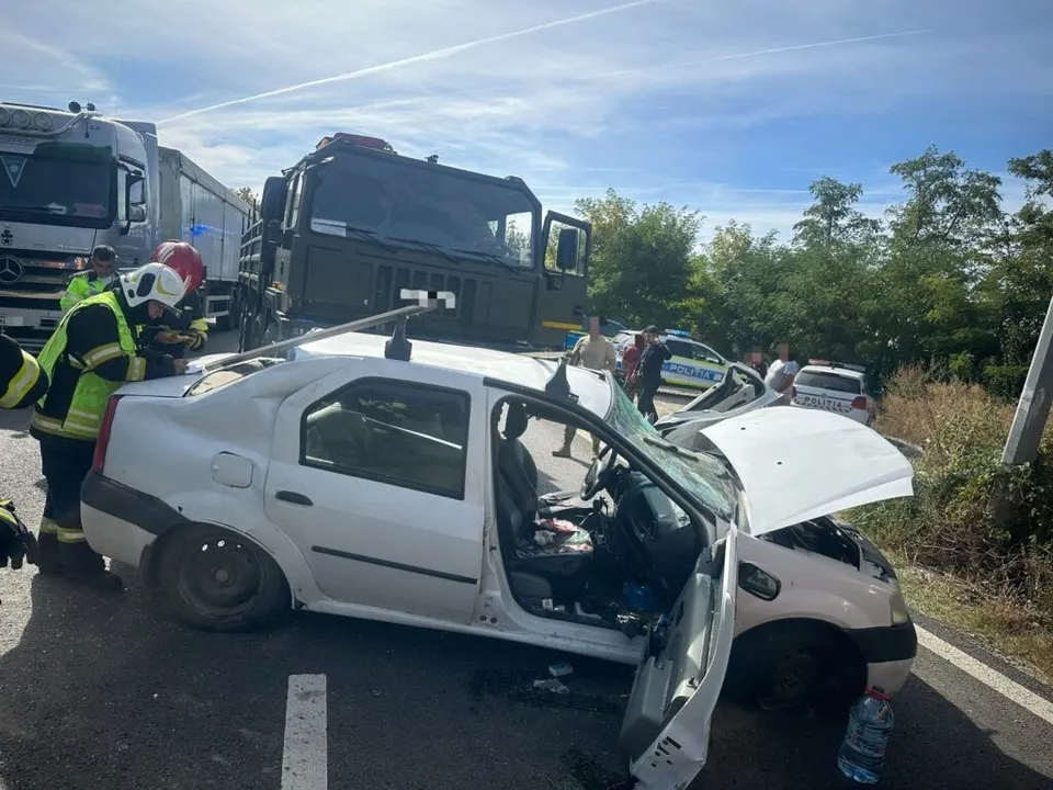 Un autoturism alb cu portierele deschise și capota ridicată stă pe asfalt lângă un camion negru și un vehicul de poliție.