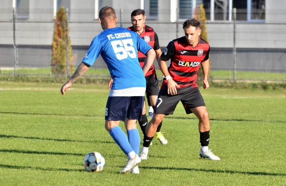 Trei jucători de fotbal sunt pe un teren de gazon verde, unul având un balon la picioare și purtând un echipament albastru.