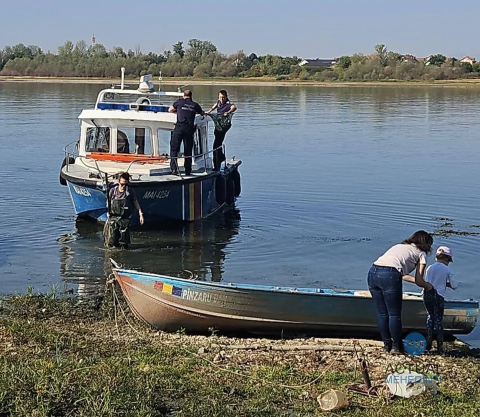 Trei persoane stau lângă o barcă mică pe malul unui râu, în timp ce alți doi oameni se află pe un vas motorizat mai mare.
