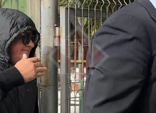Un bărbat cu o jachetă neagră cu glugă și ochelari de soare stă lângă un stâlp metalic și o poartă de sârmă.