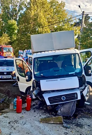 Un vehicul alb cu compartiment de marfă este oprit pe un drum, având partea frontală deteriorată și ușa șoferului deschisă.
