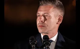 Un bărbat cu părul scurt și coafat în sus poartă un costum închis și stă în fața a două microfoane negre.