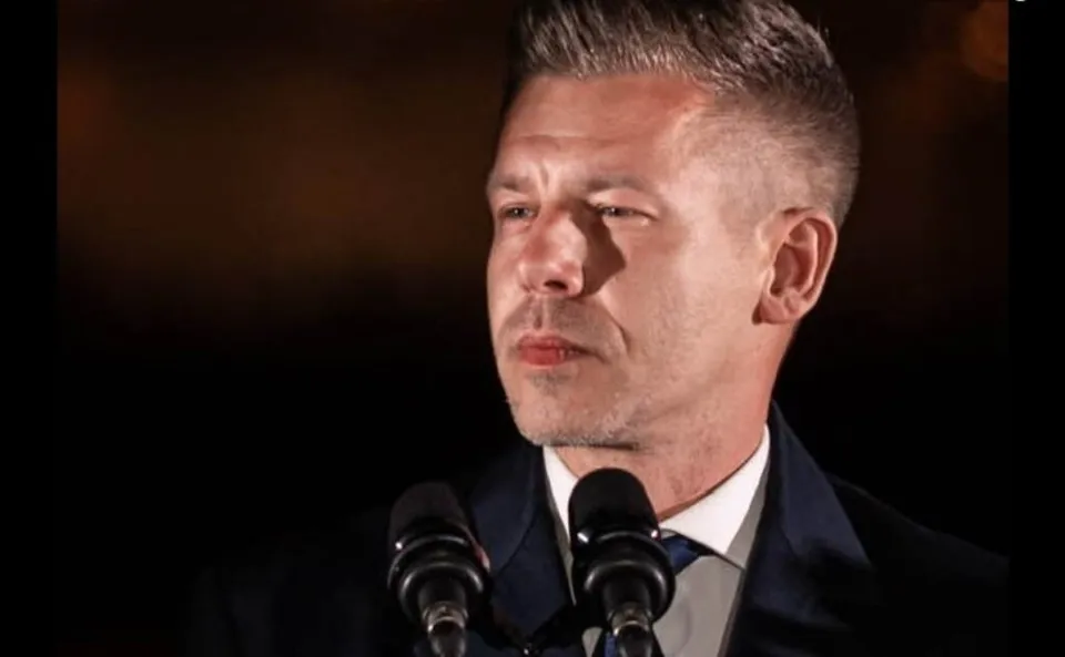 Un bărbat cu părul scurt și coafat în sus poartă un costum închis și stă în fața a două microfoane negre.