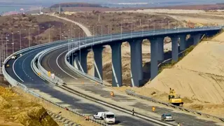 O autostradă cu mai multe benzi de rulare parcurge un viaduct înalt susținut de piloni gri într-un peisaj de dealuri deschise.