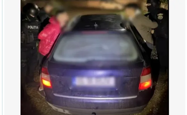 O mașină de culoare închisă este înconjurată de mai mulți oameni, dintre care unii poartă uniformă neagră cu inscripția POLITIA.