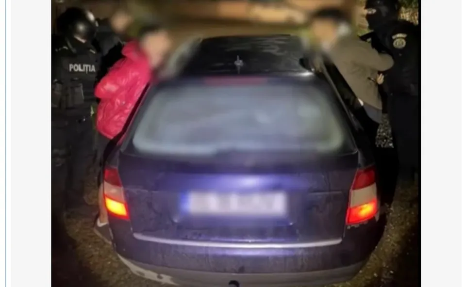 O mașină de culoare închisă este înconjurată de mai mulți oameni, dintre care unii poartă uniformă neagră cu inscripția POLITIA.