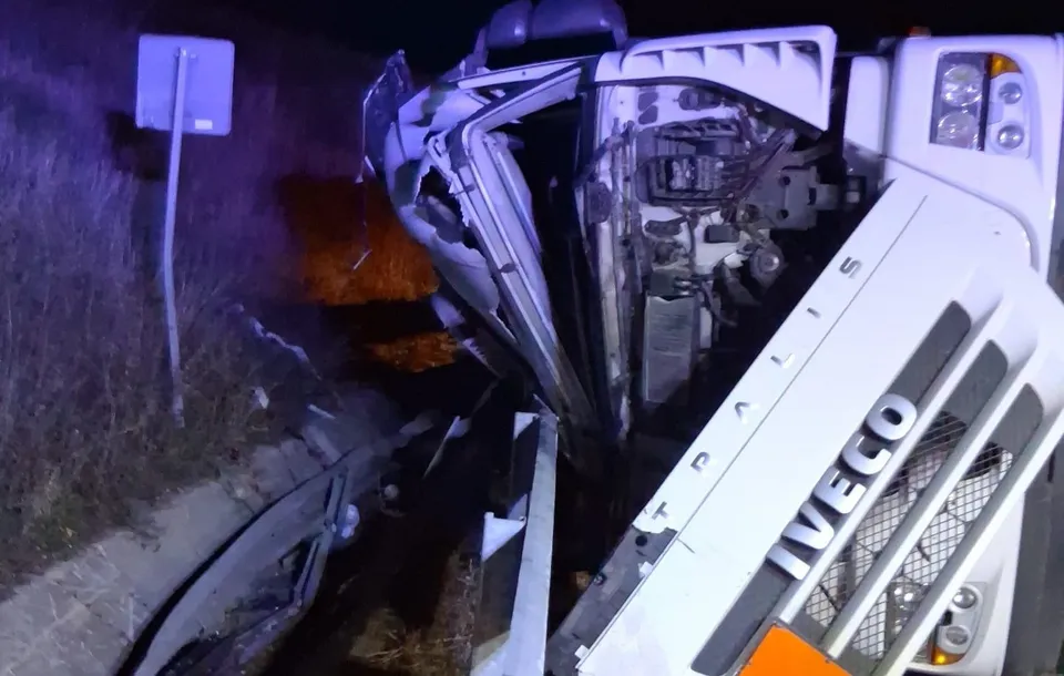 Un camion alb de marca Iveco este răsturnat pe o laterală, într-un mediu întunecat cu vegetație în fundal.