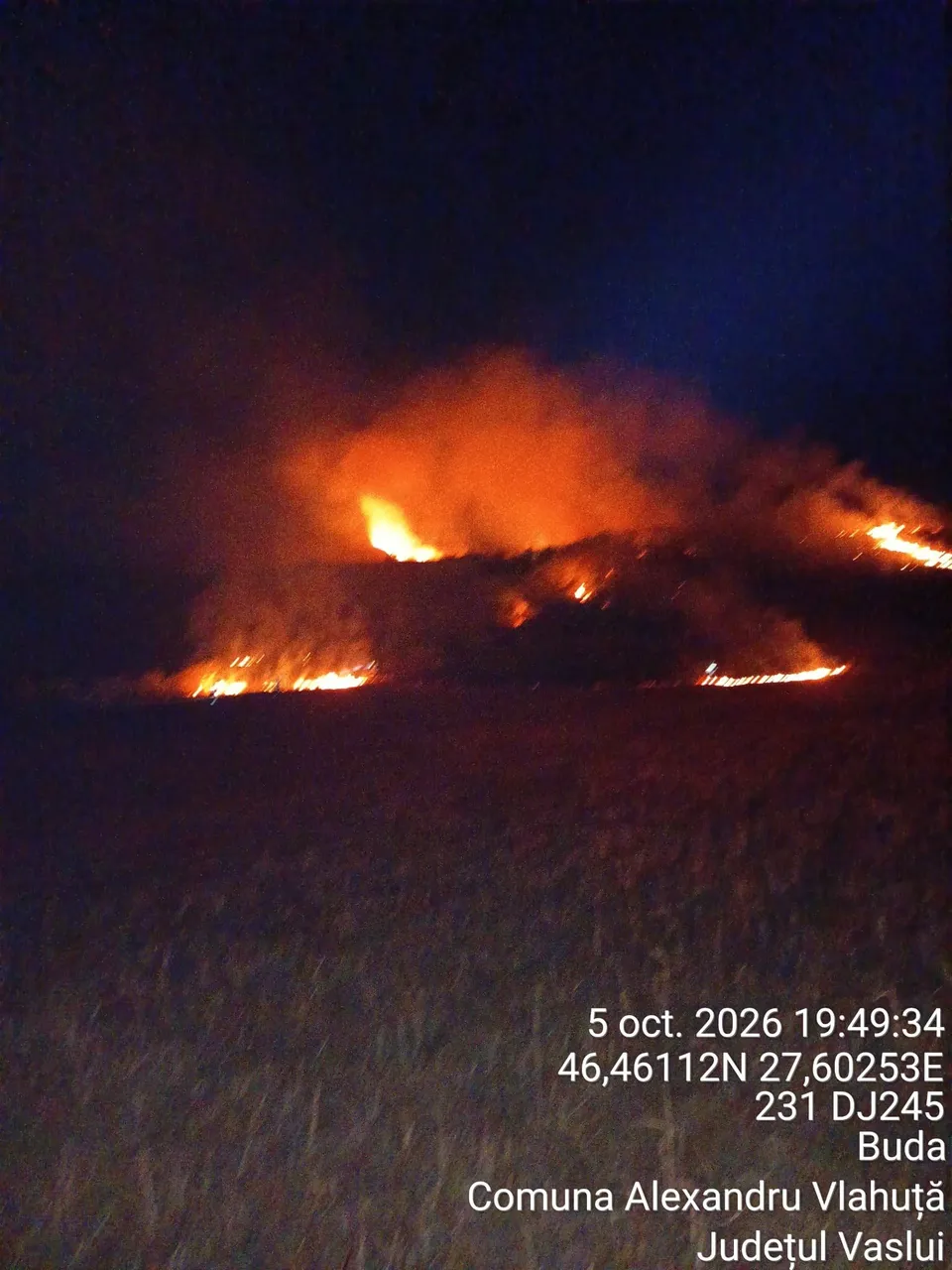 Flăcări și fum portocaliu se ridică deasupra unui teren întunecat sub un cer de noapte.