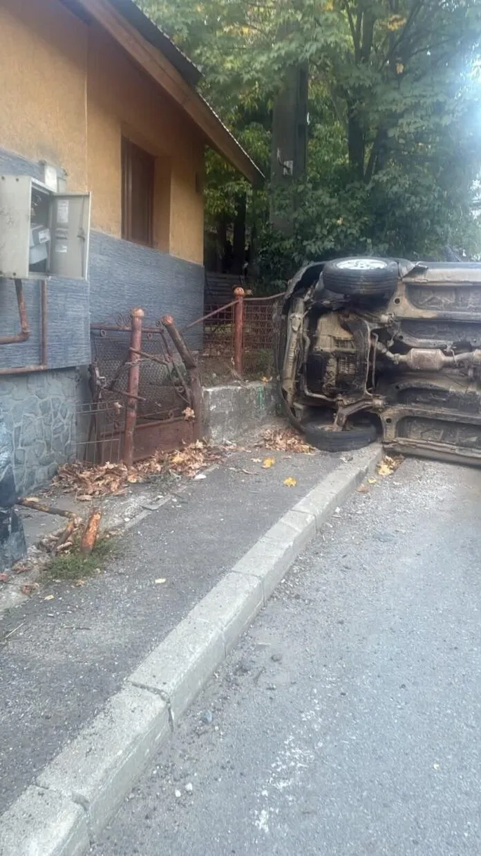 Un autoturism răsturnat se află pe o zonă de trotuar lângă o clădire și un gard metalic.
