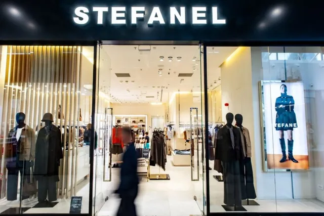 Fațada unui magazin de haine cu manechine expuse în vitrine și un semn luminos cu textul STEFANEL deasupra intrării.