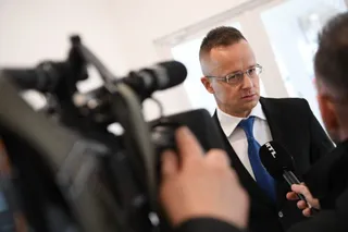 Un bărbat cu ochelari și cravată albastră este încadrat de microfoane și camere de filmare într-un cadru interior.