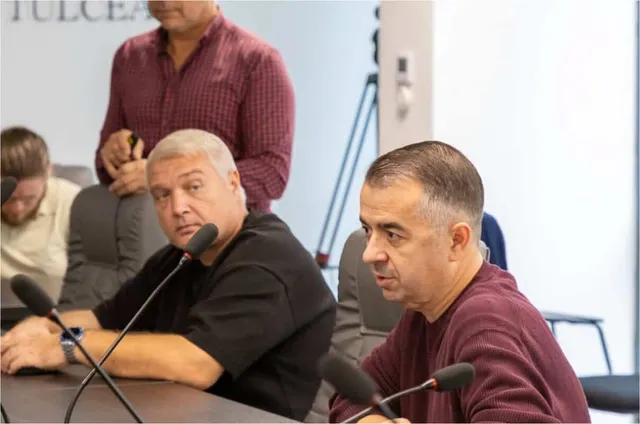 Trei bărbați stau la o masă cu microfoane, unul purtând un tricou negru și celălalt un tricou bordo.
