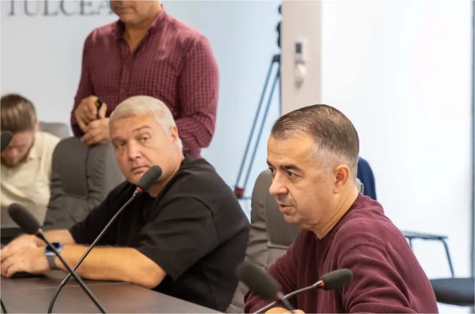 Trei bărbați stau la o masă cu microfoane, unul purtând un tricou negru și celălalt un tricou bordo.