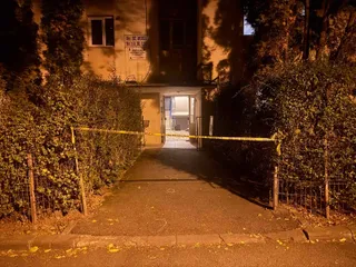 Un cordon de poliție galben este întins pe un drum pietonal care duce spre intrarea unei clădiri înconjurate de vegetație densă noaptea.