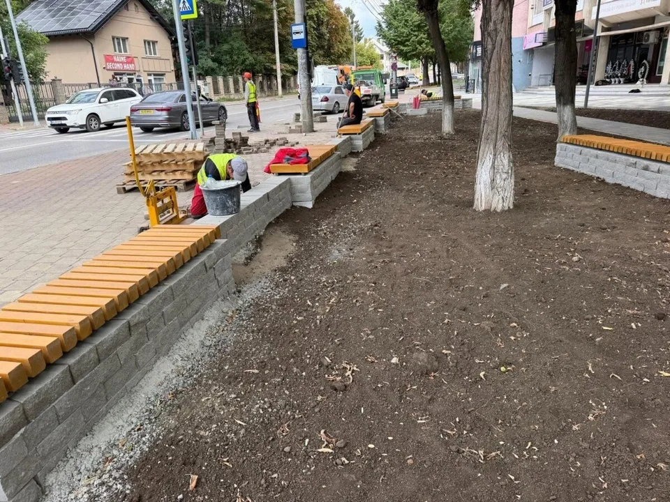 Imaginea prezintă un șantier în aer liber, cu bănci noi din lemn și beton, pământ proaspăt și muncitori. Pe fundal se observă o stradă cu mașini și clădiri.
