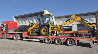 Un buldoexcavator galben este transportat pe o remorcă de tip platformă trasată de un camion roșu sub un cer senin.
