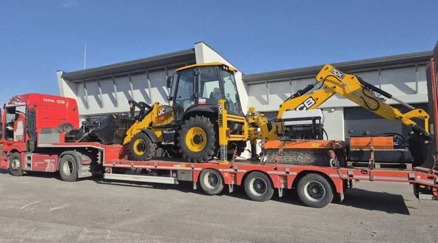 Un buldoexcavator galben este transportat pe o remorcă de tip platformă trasată de un camion roșu sub un cer senin.