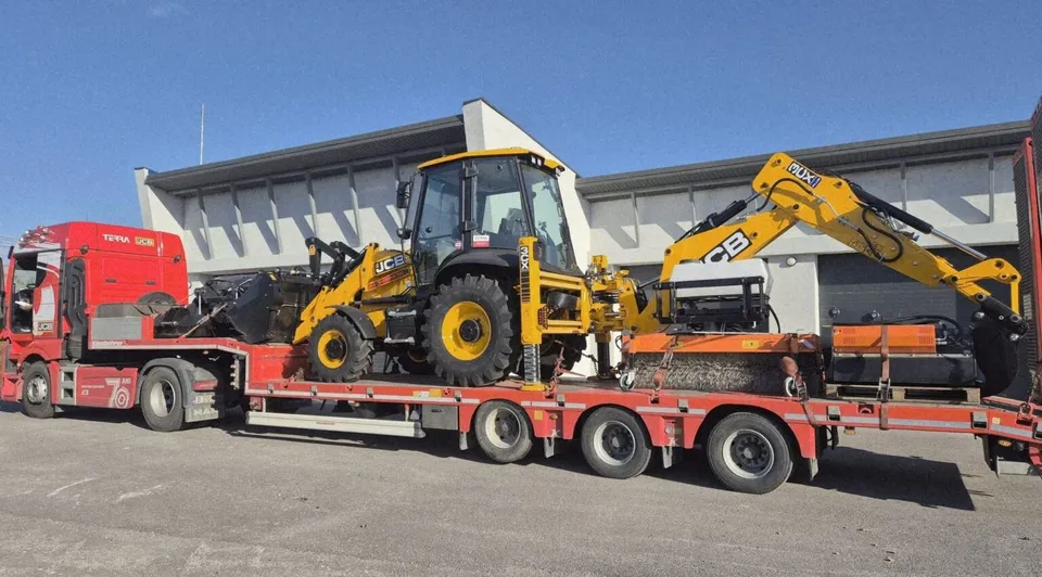 Un buldoexcavator galben este transportat pe o remorcă de tip platformă trasată de un camion roșu sub un cer senin.