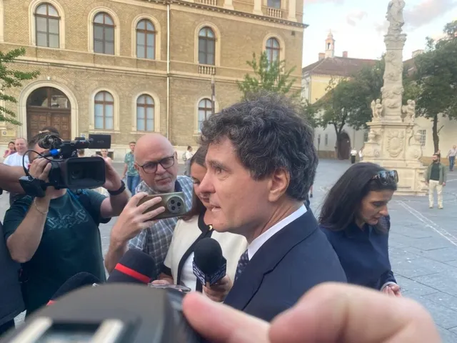 Un bărbat într-un costum închis la culoare vorbește în fața mai multor microfoane, în timp ce persoane cu aparate foto și telefoane îl filmeză.