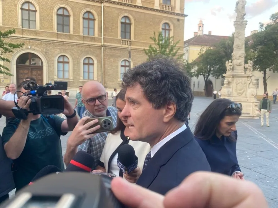 Un bărbat într-un costum închis la culoare vorbește în fața mai multor microfoane, în timp ce persoane cu aparate foto și telefoane îl filmeză.
