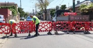 Două persoane purtând veste fluorescente de siguranță lucrează pe un drum delimitat de bariere de plastic roșu și alb lângă un camion.