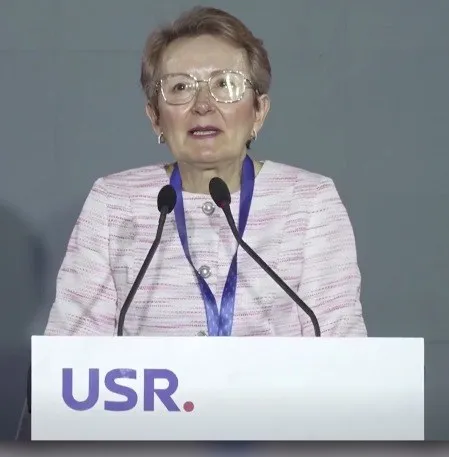O femeie cu ochelari și sacou roz deschis stă în spatele unui podium alb pe care este scris textul USR.