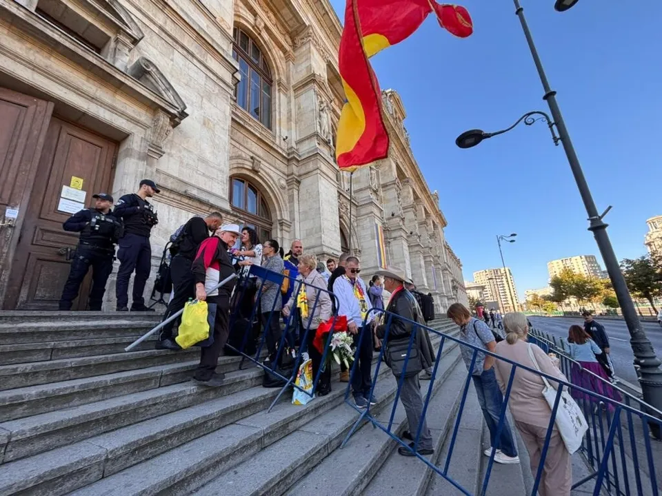 O grupă de oameni stă pe scările unei clădiri înalte, lângă o barieră metalică albastră, sub un steag roșu și galben.