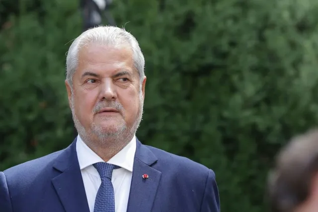 Un bărbat cu părul alb și barbă scurtă poartă un costum albastru, cămașă albă și cravată albastră în fața unui fundal verde de vegetație.