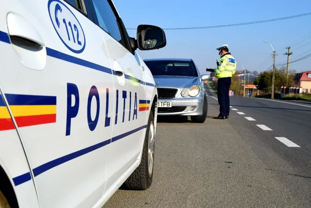 O mașină de poliție albă cu inscripții albastre stă pe marginea unui drum lângă un vehicul gri și un polițist în uniformă galbenă.