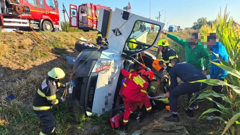 Echipe de salvatori în uniforme colorate lucrează lângă o autoutilitară albă răsturnată într-un șanț, în prezența unui camion de pompieri roșu.