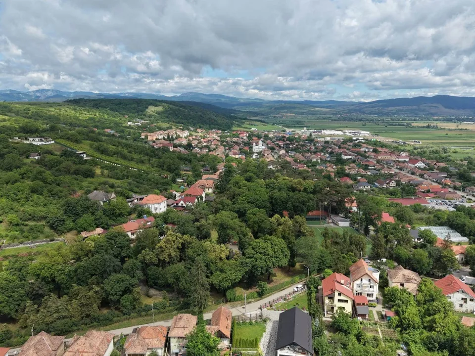 O vedere aeriană asupra unui oraș cu case cu acoperișuri roșii, dealuri verzi și un cer înconjurat de nori.