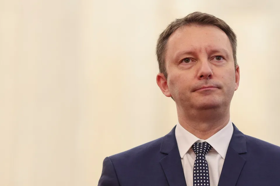 Un bărbat cu părul scurt, purtând sacou albastru, cămașă albă și cravată cu puncte albe, privește spre lateral pe un fundal deschis la culoare.