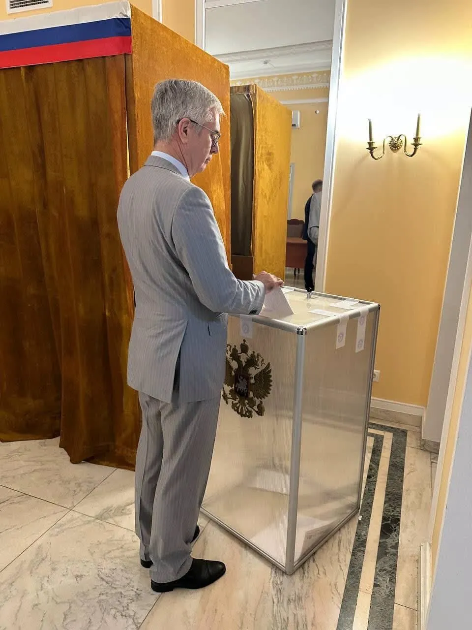 Un bărbat cu ochelari și costum gri introduce un plic într-o urnă transparentă situată într-o încăpere cu pereți galbeni și draperii maronii.