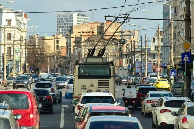 Un trolebus alb circulă pe un drum aglomerat cu numeroase mașini colorate sub un cer senin, în fața unor clădiri urbane.