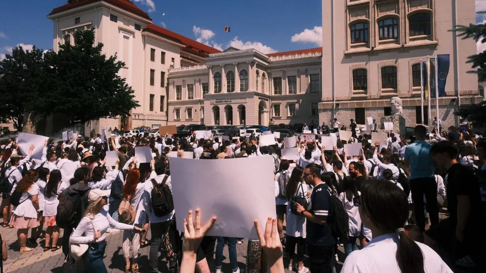 O mulțime de persoane adunată într-o piață publică din fața unei clădiri universitare, ținând panouri albe de protest.