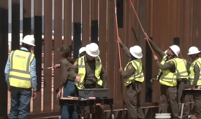 Mai mulți muncitori cu căști albe și veste reflectorizante galben-verzui lucrează lângă o structură metalică formată din stâlpi verticali maro.