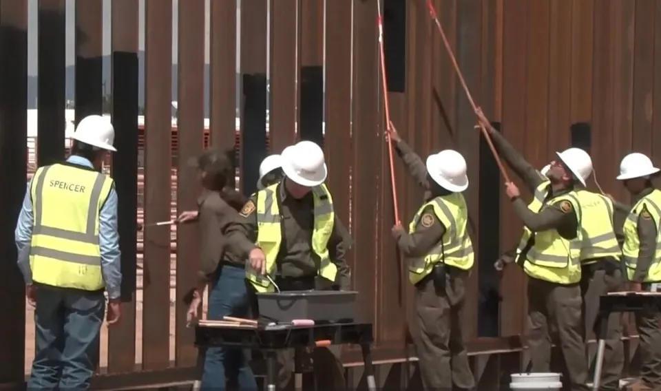 Mai mulți muncitori cu căști albe și veste reflectorizante galben-verzui lucrează lângă o structură metalică formată din stâlpi verticali maro.