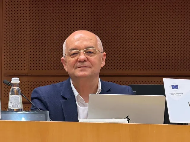 Un bărbat cu ochelari și sacou albastru stă la o masă lângă un laptop și o sticlă de apă în fața unui perete perforat maro.