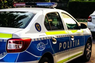 Un autovehicul de poliție alb cu marcaje albastre și galbene este parcat pe un drum lângă vegetație.