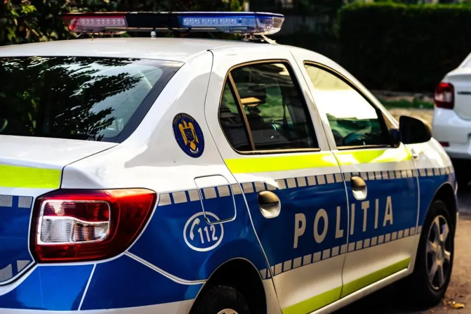 Un autovehicul de poliție alb cu marcaje albastre și galbene este parcat pe un drum lângă vegetație.