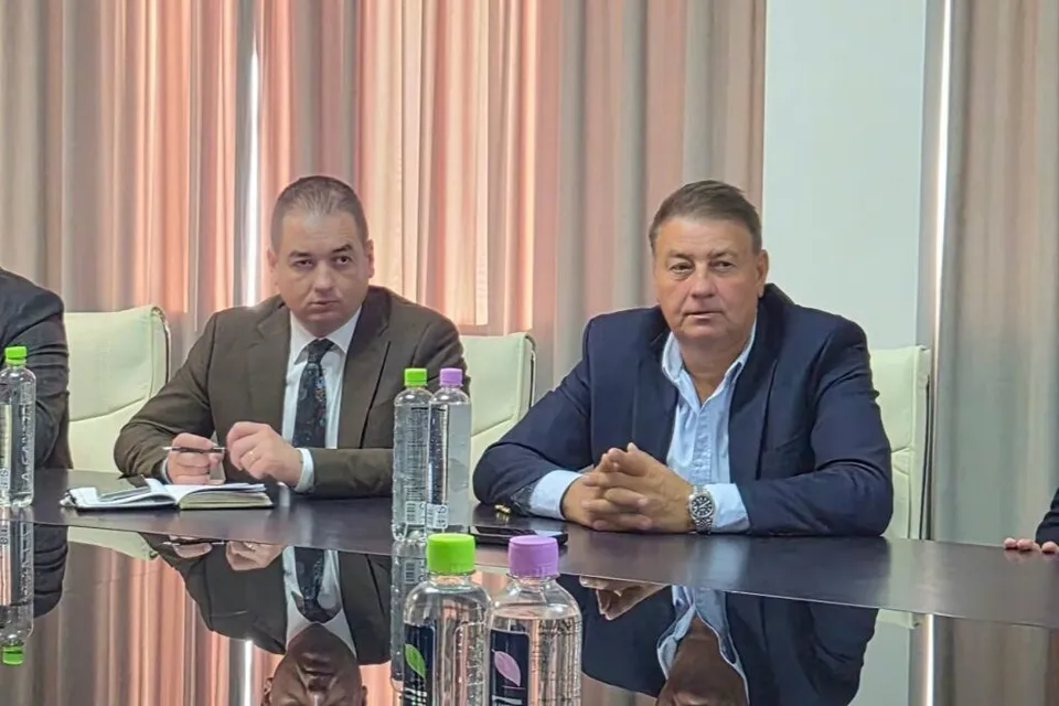 Doi bărbați stau la o masă de conferințe, unul purtând un sacou maro cu cravată și celălalt un sacou albastru cu cămașă deschisă.