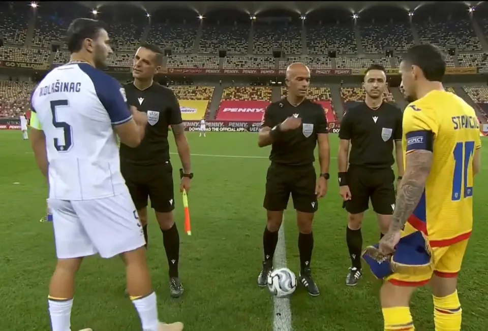 Patru bărbați în echipamente negre stau pe un teren de fotbal lângă un jucător în echipament alb și unul în echipament galben.