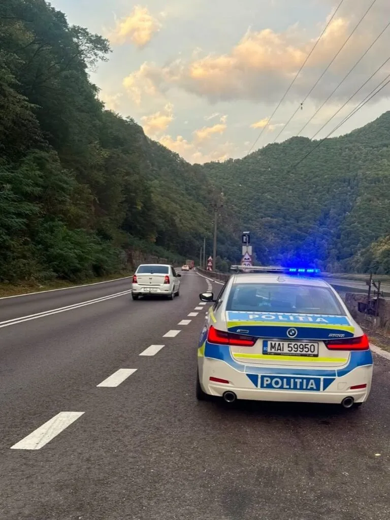 O mașină de poliție albă cu lumini albastre aprinse circulă pe un drum asfaltat, în fața unei mașini albe, într-o zonă montană cu vegetație.