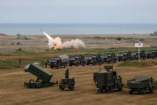 Un lansator înclinat aflat în plan apropiat, vehicule militare aliniate, o rachetă în zbor și fum pe un teren deschis.