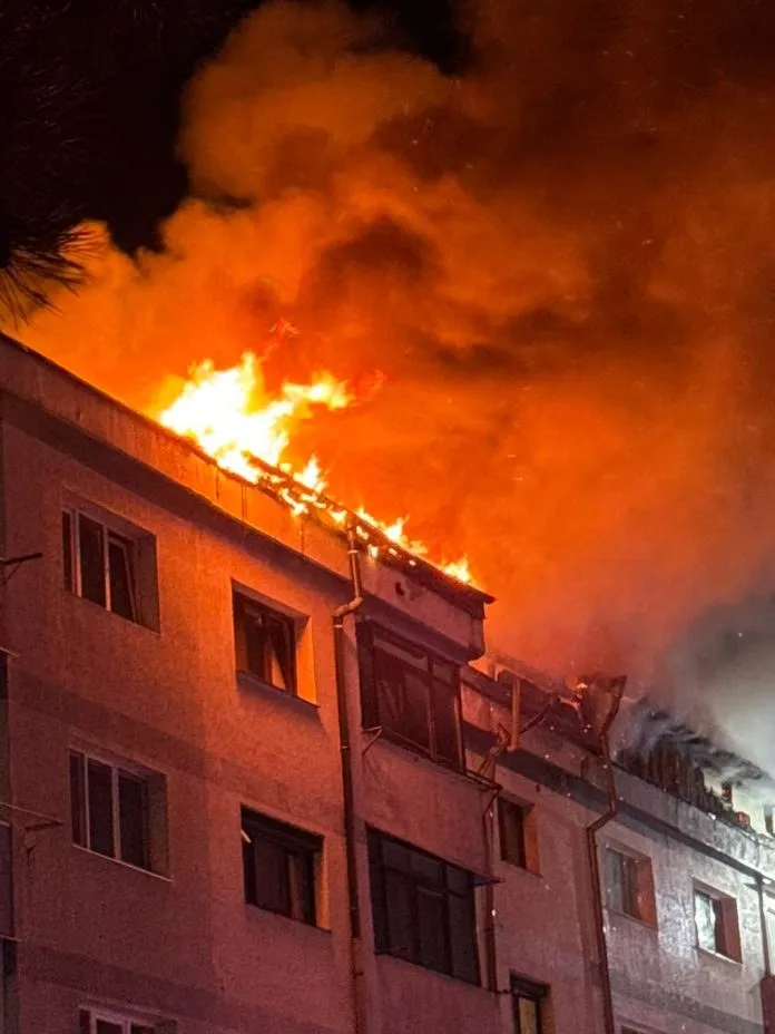 Flăcări și fum dens se ridică de pe acoperișul unei clădiri de locuințe pe timpul nopții.