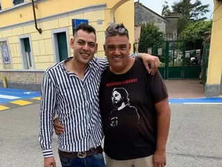 Doi bărbați stau unul lângă altul, unul purtând o cămașă cu dungi verticale și celălalt un tricou negru cu o reprezentare grafică pe piept.