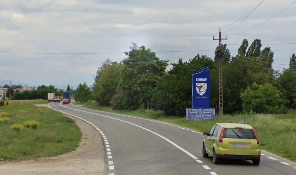 O drumuită asfaltată curbează spre stânga, cu o mașină galbenă în prim plan și un panou albastru de bun venit la Ghimbav în fundal.