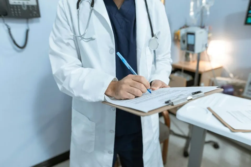 Un medic într-un halat alb și echipament medical scrie pe o foaie de hârtie fixată pe o clipboard.