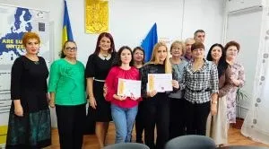Un grup de femei stă în picioare într-o încăpere, unele purtând documente albe în mâini, în fața unor steaguri și a unui panou informativ.